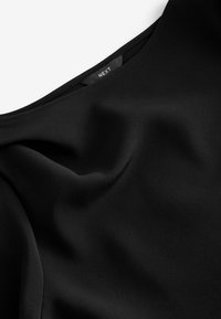 Haut en tissu noir avec une texture lisse et un décolleté large, présentant un léger éclat et un bord cousu. Étiquette de marque visible à l'intérieur.