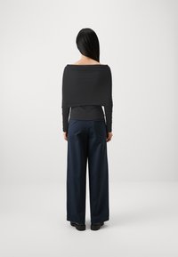 Zwarte top met lange mouwen en off-shoulder ontwerp, gecombineerd met navy wijde broek en zwarte chunky schoenen, gezien vanaf de achterkant.