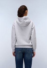 Sudadera gris con capucha, de textura suave y puños acanalados, combinada con vaqueros azules con bolsillos traseros. Vista desde la parte trasera.