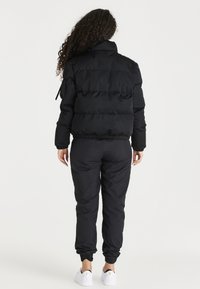 Svart pufferjacka med hög krage och ribbad nederkant, tillsammans med svarta smala byxor. Vita sneakers med en guldlogo som accent.