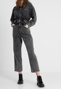 Czarna koszula z denimu z zapięciem na guziki i dwoma kieszeniami na piersiach, w połączeniu z czarnymi dżinsami z denimu o wysokim stanie, prostym kroju nogawki i podwiniętym mankietem. Dopełnieniem stroju są czarne botki za kostkę sznurowane.