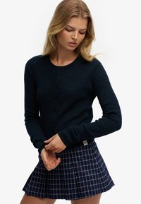 Marineblå strikket cardigan med ribbet struktur og knapper foran. Kombineret med et ternet plisseret skørt i marineblå, rød og hvid.