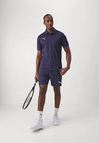 Marineblå poloshirt og shorts, begge laget av pustende stoff, med hvite Puma-logo detaljer. Utstyrt med en tenniskølle og hvite sneakers.