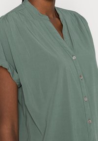 Grön blus med kort ärm gjord av mjukt tyg, med ståkrage och knapprad framtill. Slät textur med rynkade detaljer.