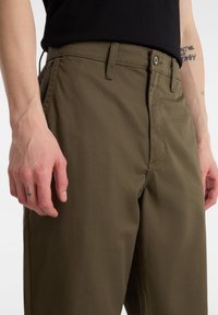 Pantaloni verde oliva realizzati in un tessuto resistente. Presentano una parte frontale piatta con chiusura a zip, passanti per la cintura e una tasca laterale visibile.