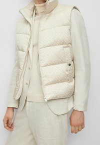 Lightweight, beige puffer vest with a high collar. Patterned texture, zip closure, and side pockets. Worn over a cream-colored knit top.