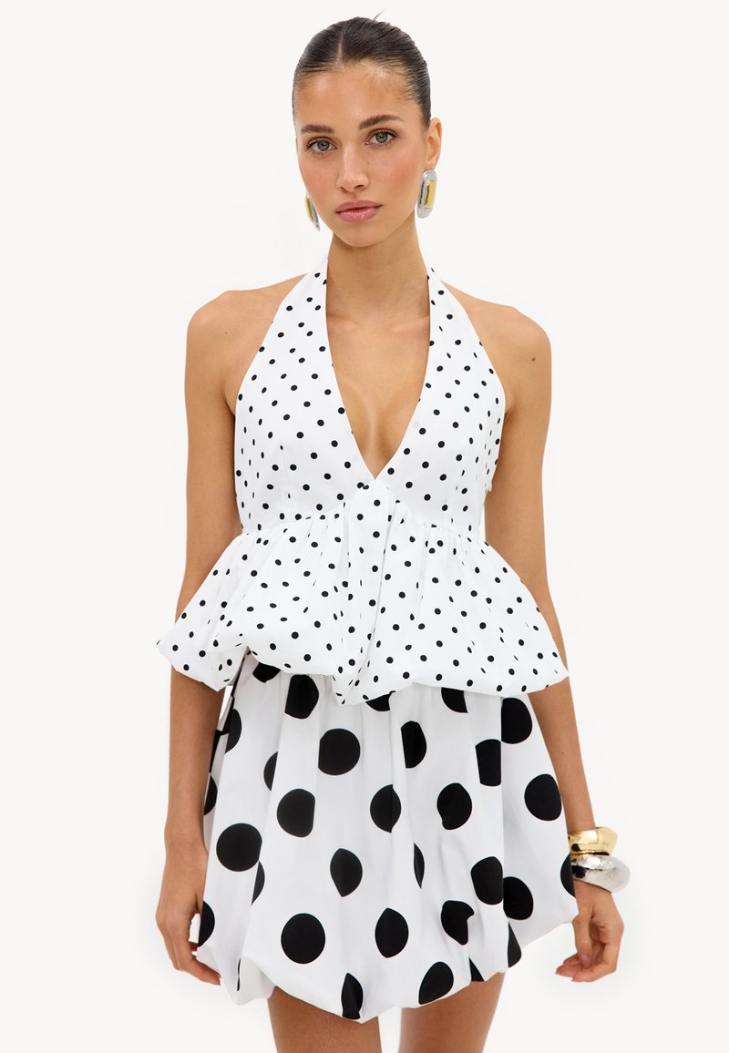 Femme portant un haut blanc à col halter avec de petits pois noirs et une jupe assortie avec de gros pois noirs, accessoirisée de bracelets.
