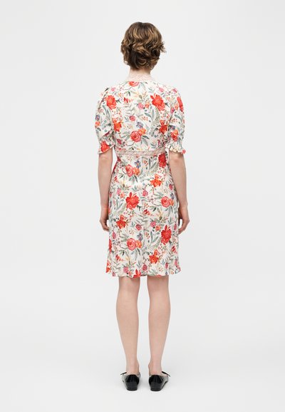 Γυναίκα με κοντά μαλλιά, που φοράει λευκό φόρεμα μήκους έως το γόνατο με κόκκινα και ροζ λουλουδάτα σχέδια και μαύρα επίπεδα παπούτσια, όρθια με την πλάτη προς τα εμπρός.