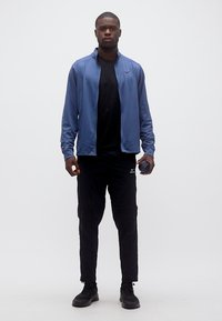 Νέος άνδρας που στέκεται, φορώντας μπλε μπουφάν με φερμουάρ, μαύρο πουκάμισο και παντελόνι, κρατώντας ένα μικρό μαύρο κυλινδρικό αντικείμενο στο δεξί χέρι.