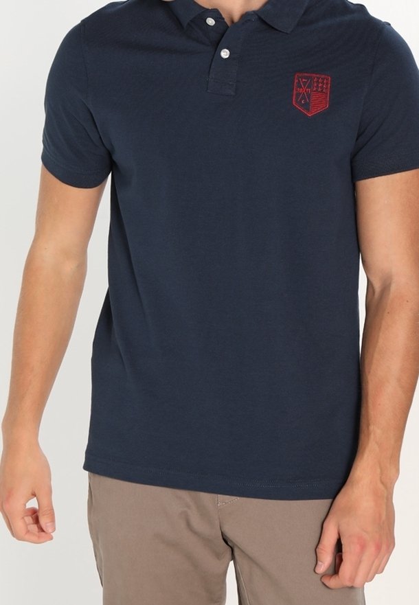 Homme portant un polo bleu marine à manches courtes avec un emblème brodé rouge et un pantalon beige, debout devant un fond uni.