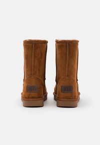 Hnědé nubukové kotníkové boty s měkkou texturou, s plochou podrážkou a malou logovou nášivkou na zadní straně. Jednoduchý design, bez vzorů nebo ozdob.