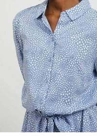 Ljusblå knapp-up skjorta med vita prickar, med krage och en samlad nederkant med knyt, tillverkad av ett mjukt tyg.