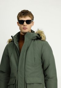 Młody mężczyzna w czarnych okularach przeciwsłonecznych, zielonej zimowej parce z kapturem wykończonym futrem i brązowym swetrze z golfem, na jednolitym jasnym tle.