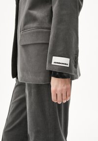 Grijze corduroy blazer met een subtiele ribbeltextuur, drie knopen op de mouw en een merklabel met "ARMEDANGELS" in het zwart.