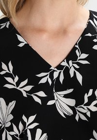 Tessuto nero con motivo floreale bianco, presenta una scollatura a V. Foglie e fiori sono contornati da forme dettagliate e una texture liscia.