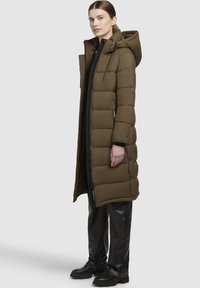 Lang, oliven grøn pufferjakke med hætte, der har vandrette syninger, sidelommer, en fuld lynlås og en afslappet pasform. Kombineret med sorte bukser.