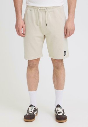 Man draagt beige shorts met trekkoord, zwarte en witte sneakers en witte sportsokken, staand tegen een witte achtergrond.