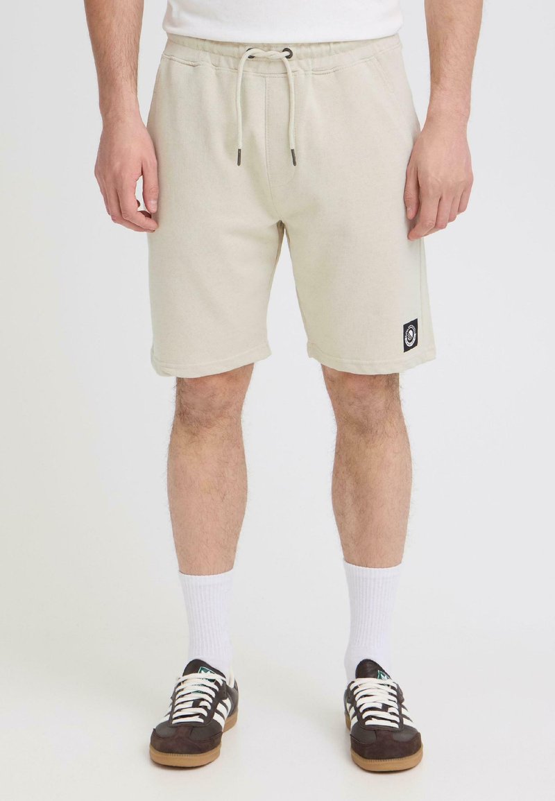 Homme portant un short beige à cordon de serrage, des baskets noires et blanches, et des chaussettes blanches montant au mollet, debout devant un fond blanc.