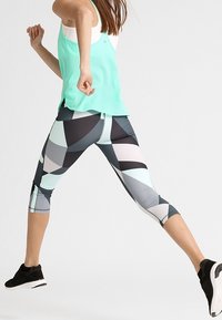 Kolorowe legginsy sportowe z geometrycznymi wzorami w czerni, szarości, jasnoniebieskim i różowym; połączone z miętowym topem na ramiączkach i czarnymi sneakersami.