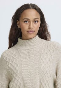 Maglione a maglia beige con collo alto, caratterizzato da distintivi motivi a treccia da un lato e una texture liscia dall'altro.