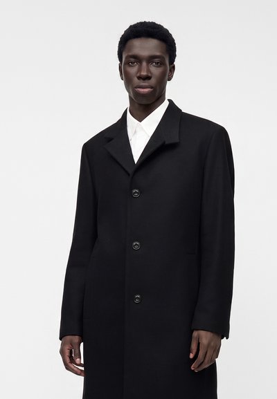 Jeune homme noir portant un manteau noir boutonné sur une chemise blanche, debout devant un fond gris clair uni.