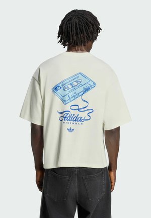 Mann mit dunklem lockigem Haar trägt weißes übergroßes T-Shirt mit blauem Kassettenband und Adidas Originals Logo auf dem Rücken.