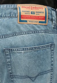 Világoskék farmernadrág barna bőr címkével a derékrészen. A címkén kék és piros szöveg található, amely márkára és minőségre vonatkozó részleteket tartalmaz.