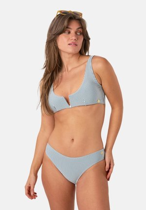 Vrouw draagt een blauw-wit gestreepte bikini met een V-hals top en hoog uitgesneden broekje, kijkt naar links met een zonnebril op haar hoofd.
