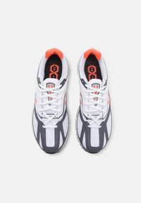 Une paire de chaussures de sport blanches et grises avec des accents et des lacets orange, vue de dessus sur un fond uni.