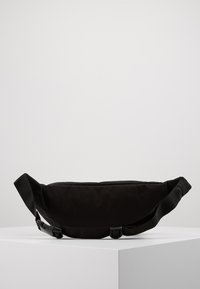 Bolso bandolera negro hecho de tela texturizada, con forma curva, correa ajustable y cierre de hebilla, exhibido sobre una superficie blanca.