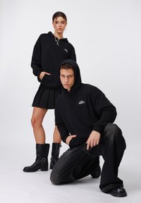 Sudaderas con capucha negras con acentos gráficos, combinadas con una falda plisada y botas negras hasta la rodilla; acompañadas de jeans oscuros de pierna ancha.