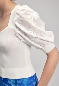Maglione bianco con corpo a maglia testurizzata e maniche a sbuffo trasparenti adornate da margherite ricamate, con orlo aderente e scollatura quadrata.