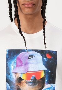 Balta marškinėliai su spalvinga grafika, vaizduojančia mešką su kibirėliu ir akiniais nuo saulės, skaitmeninėse erdvės fone su planetomis bei Nike logotipu.