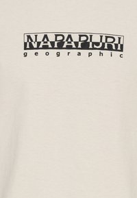 „NAPAPIJRI“ velkými černými písmeny na černém zakřiveném obdélníkovém pozadí a „geographic“ malými písmeny pod ním na béžové látce.