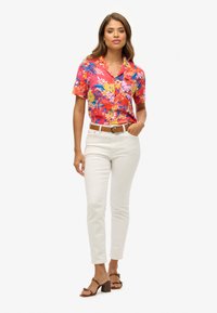 Chemise à manches courtes, rose vif à motifs floraux avec des accents bleus et jaunes, associée à un pantalon blanc slim et des sandales à talons bruns.