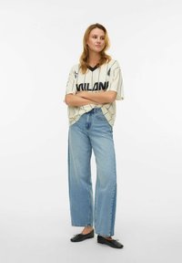 Locker sitzendes cremefarbenes T-Shirt mit schwarzen Streifen und Schriftzug, kombiniert mit hellblauen, hoch taillierten Jeans mit geradem Bein und schwarzen Ballerinas.