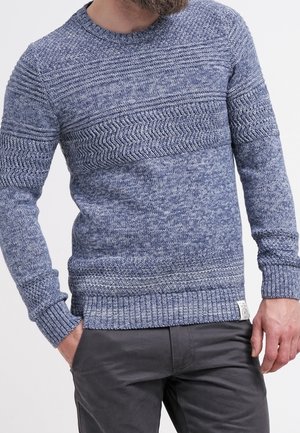 Muž v modrém pleteném svetru s texturou a vzorovanými horizontálními pruhy, doplněný tmavě šedými kalhotami, s rukama částečně v kapsách.