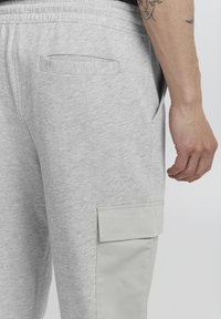 Light grey sweatpants with a smooth texture, featuring a side pocket with a flap and a back pocket. Elastic waistband visible at the top.