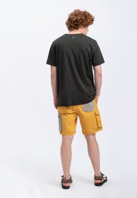 Zwarte kortemouwen t-shirt gecombineerd met gele cargoshorts met grijze accenten en zakken, gedragen met donkere sandalen. Op een schone achtergrond.