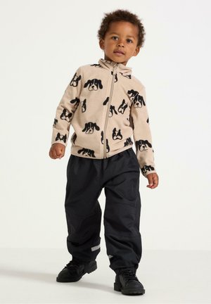 Giacca a vento beige chiaro con motivo a faccia di cane nera, abbinata a pantaloni impermeabili neri. Le scarpe sono nere, di pelle strutturata con suole in gomma.
