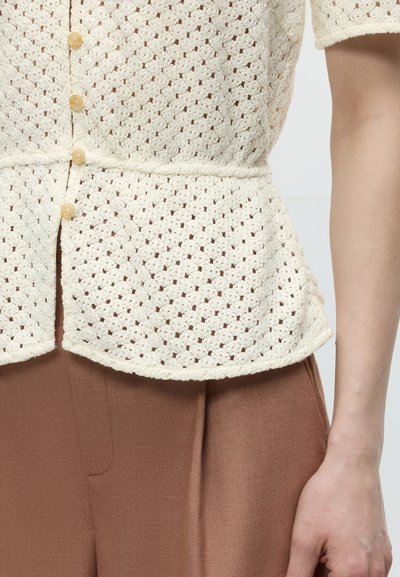 Κοντινό πλάνο ενός ατόμου που φοράει κρεμ πλεκτό τοπ με κουμπιά και καφέ παντελόνι με ψηλή μέση, δείχνοντας υφή υφάσματος και χέρι στο πλάι.