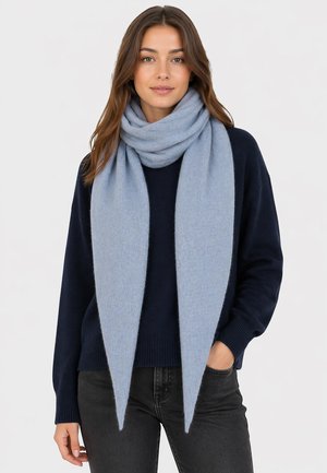 Woman wearing a light blue thick scarf wrapped around her neck, navy sweater, and dark jeans, standing with one hand in pocket.