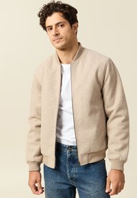 Beige fleece bomber jacket with a zip front, ribbed cuffs, and waistband. Worn over a white T-shirt, paired with blue jeans.
