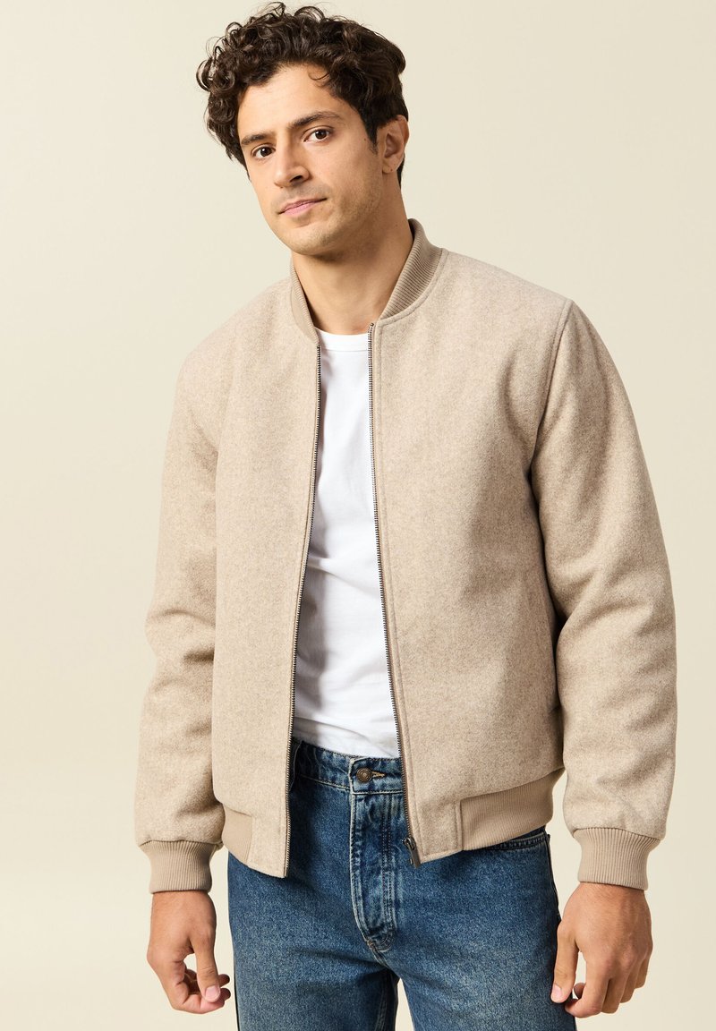 Beige fleece bomber jacket with a zip front, ribbed cuffs, and waistband. Worn over a white T-shirt, paired with blue jeans.