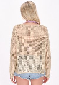 Helles beige, offenes Strickoberteil mit langen Ärmeln, oversize Passform und luftiger, lockerer Textur. Getragen über Denim-Shorts.