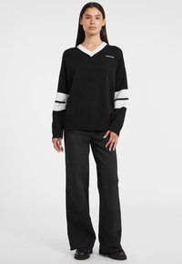 Pull noir à col en V avec des accents blancs et des rayures sur les manches, présentant un ourlet côtelé et une texture tricotée. Associé à un pantalon noir à jambes larges.