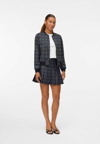 Giacca bomber a quadri nera e grigia con polsini a costine, abbinata a una mini gonna plissettata. Entrambi presentano una finitura testurizzata e una vestibilità sartoriale.