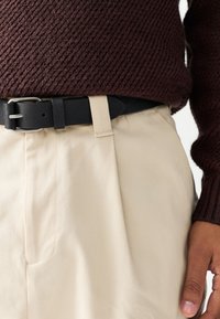 Cintura in pelle nera con fibbia in metallo argento, indossata sopra pantaloni in tessuto color crema. Maglione lavorato a maglia di colore bordeaux scuro visibile sopra.