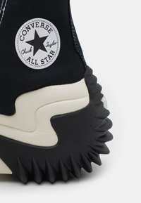 Zapatilla alta de lona negra con un logo circular blanco. Presenta una suela de goma negra gruesa con bordes dentados y acentos crema.