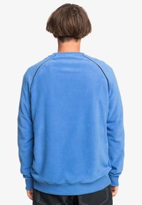 Sweatshirt en polaire bleu avec un col rond, des poignets et une taille côtelés ; dotée de passepoils foncés le long des coutures des épaules. Tissu doux et texturé.